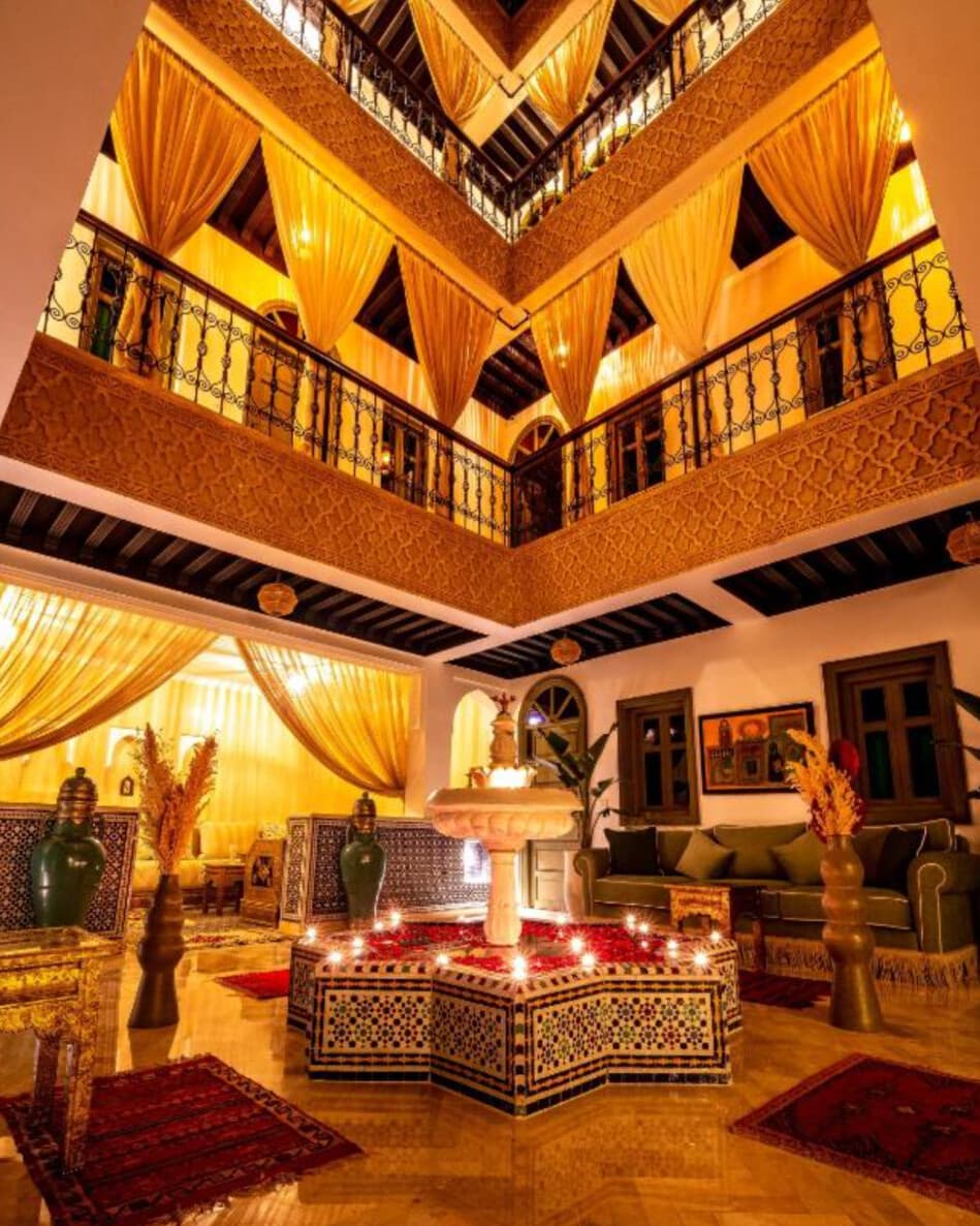 Patio central du Riad Hamdane vu depuis le rez-de-chaussée, fontaine en zellige entourée des trois étages de balcons en fer forgé et tentures dorées